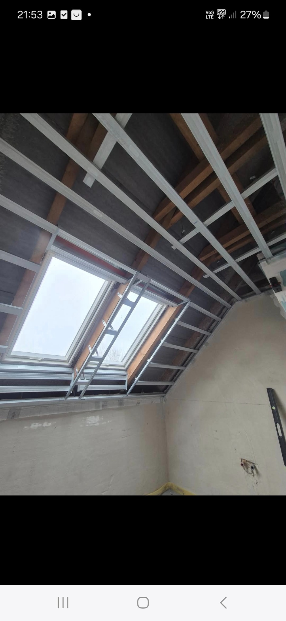 Skośny sufit z oknem dachowym w trakcie montażu stelaża z profili metalowych. Widoczne drewniane elementy konstrukcyjne dachu i fragment ściany z nierówną powierzchnią.