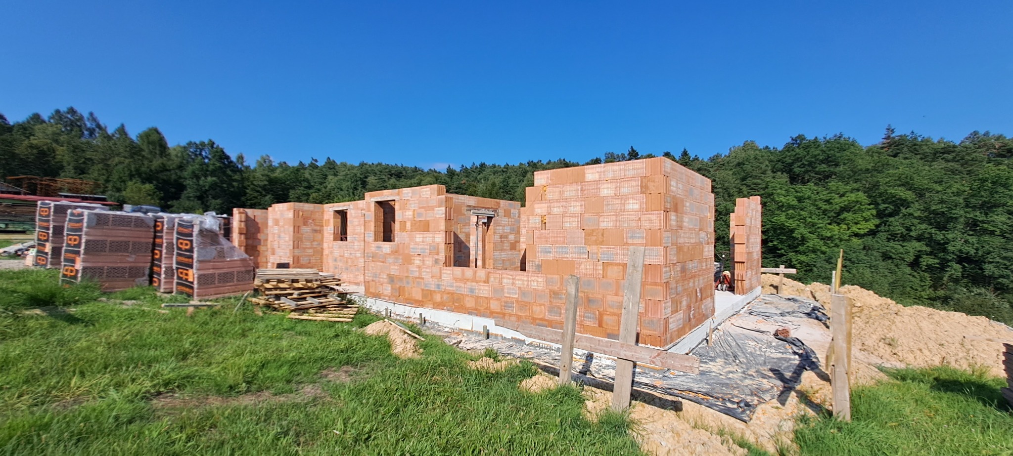 Wznoszone ściany budynku z czerwonej cegły na fundamencie, widoczne palety z cegłami i otaczający krajobraz z zielenią i drzewami w tle.