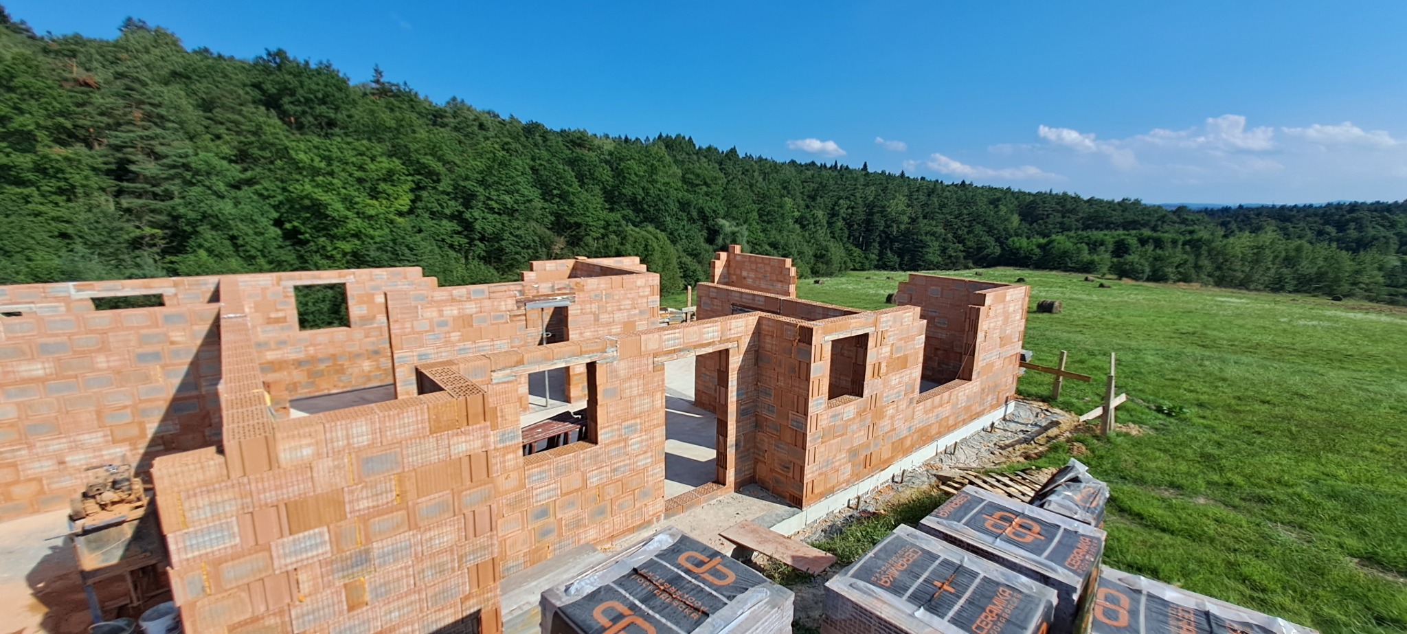 Surowy stan budynku jednorodzinnego z czerwonej cegły, widok z góry na tle zielonego lasu i błękitnego nieba, palety z materiałem budowlanym w prawym dolnym rogu.