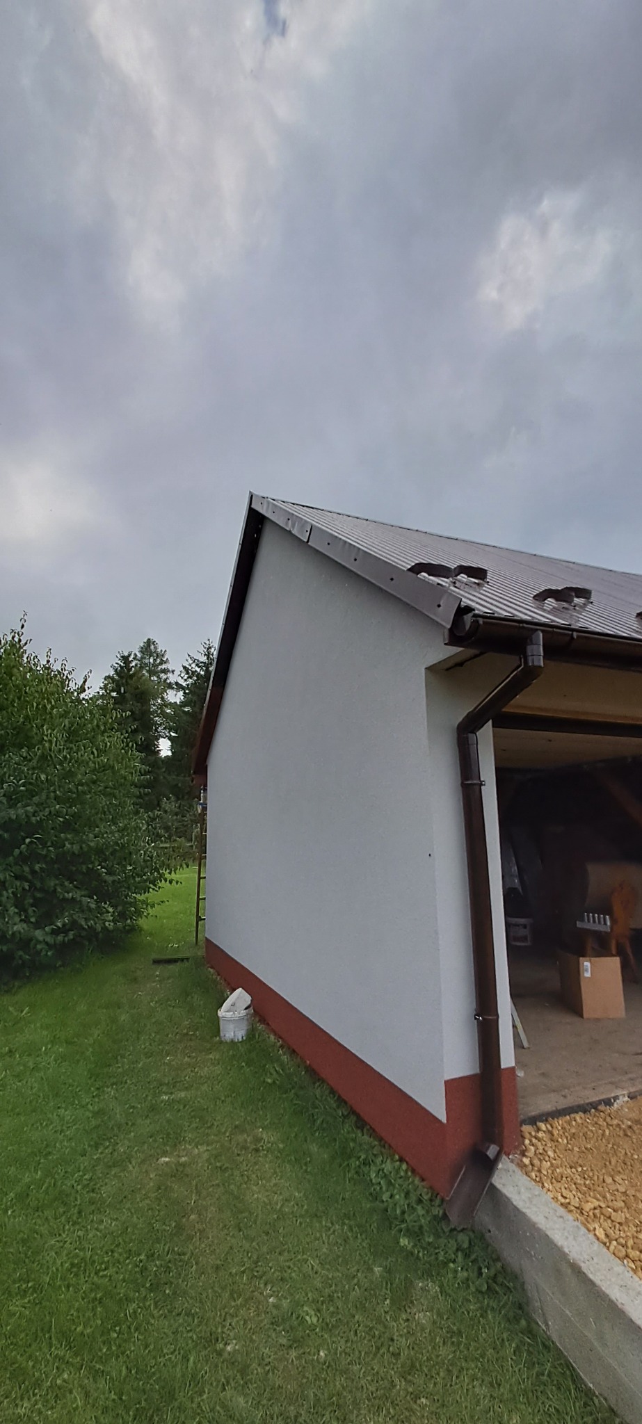 Boczny widok budynku z szarą elewacją i brązowym dachem, z widoczną rynną, fragmentem trawnika i wiadrem z farbą przy ścianie. Częściowo otwarty garaż z widocznymi przedmiotami w środku.