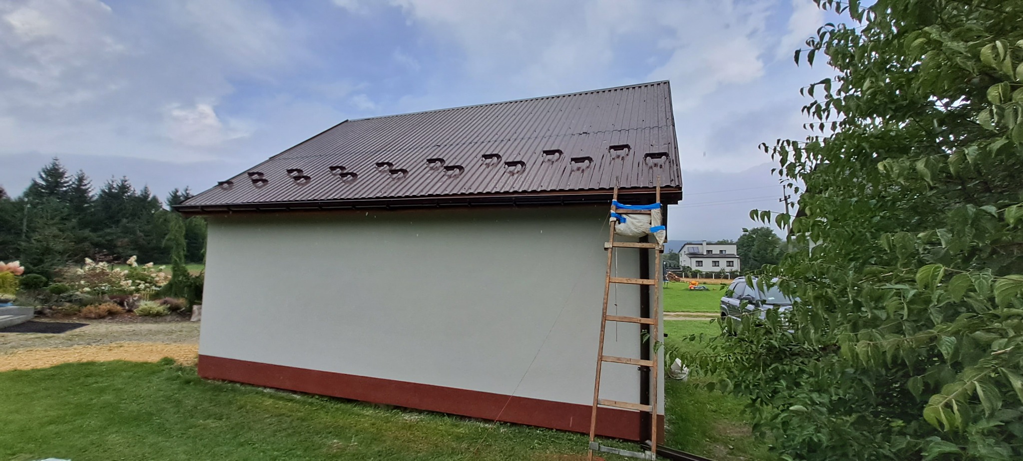 Ściana budynku z brązowym dachem z blachy trapezowej, drewniana drabina oparta o ścianę, widoczne otoczenie z zielenią i drzewami, dzień pochmurny.