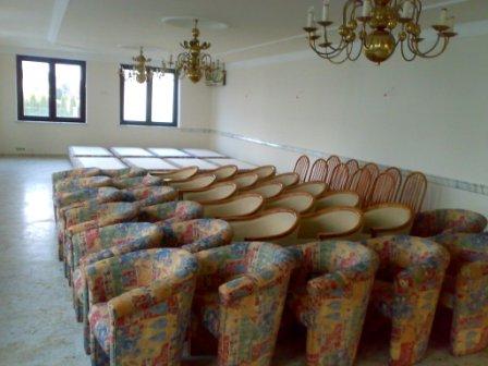 Sala z rzędami krzeseł i foteli w kolorowej tapicerce, przygotowana do prania, widoczne dwa żyrandole w tle.