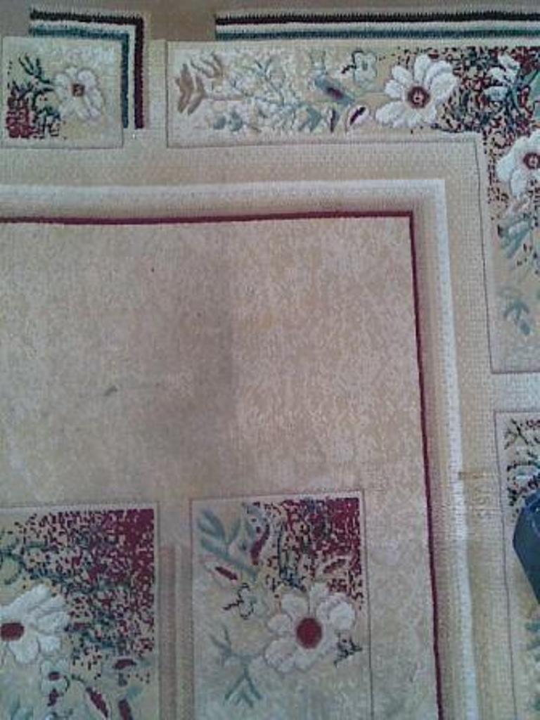 Fragment beżowego dywanu z kwiatowym wzorem i bordową obwódką, widoczny z bliska, z elementami dekoracyjnymi w rogach i na brzegach.