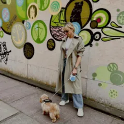 Kobieta w beżowym płaszczu i okularach przeciwsłonecznych spaceruje z psem na smyczy na tle ściany z kolorowymi graffiti.