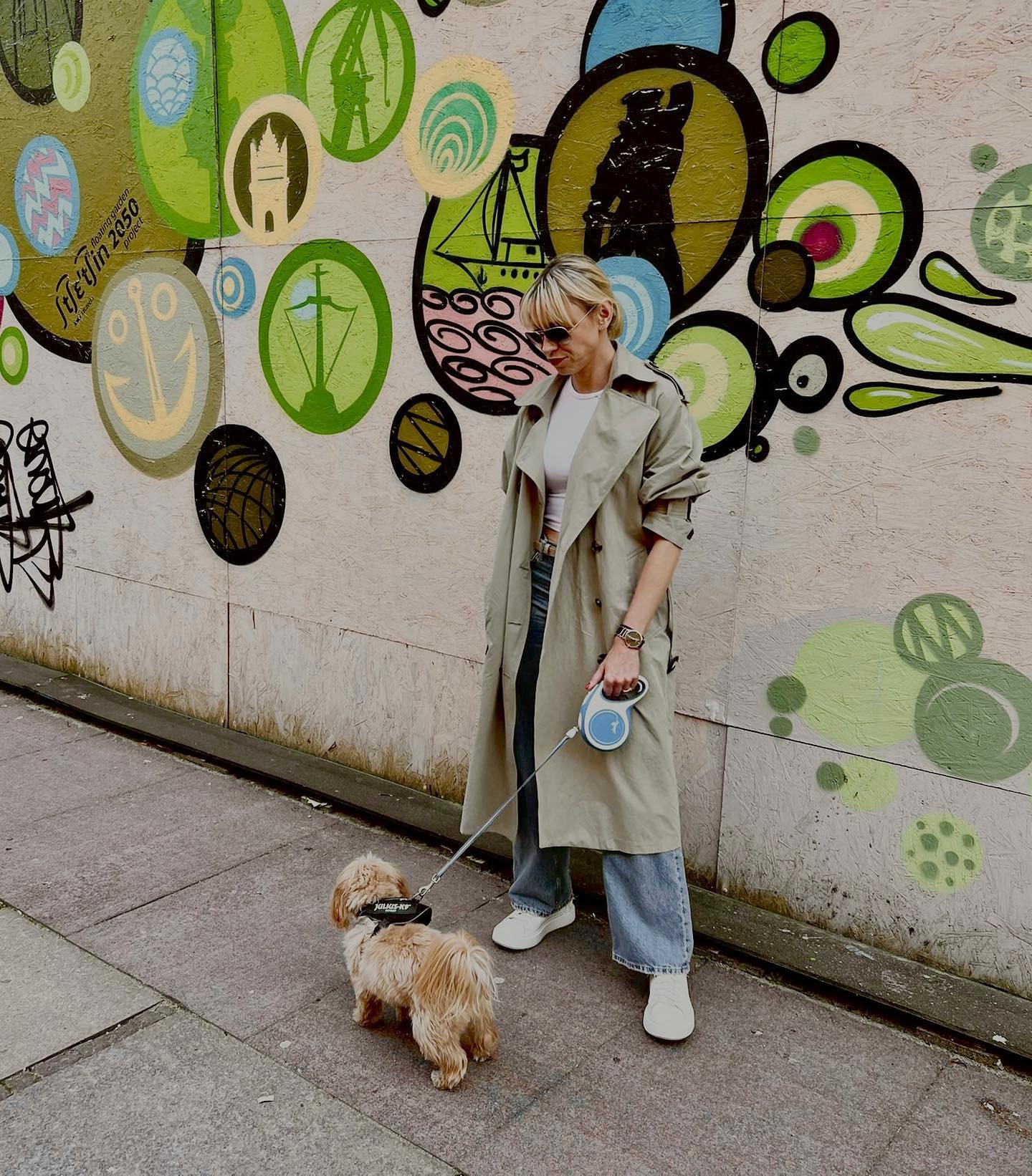 Kobieta w beżowym płaszczu i okularach przeciwsłonecznych spaceruje z psem na smyczy na tle ściany z kolorowymi graffiti.