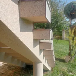 Zewnętrzne schody betonowe z wykończeniem typu baranek, prowadzące do budynku, widoczne z boku, w tle trawnik i ogrodzenie.