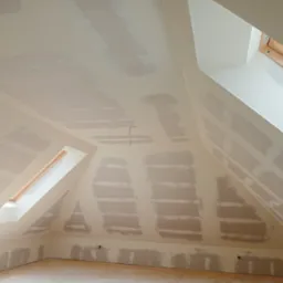 Poddasze ze skośnymi ścianami i oknami dachowymi w trakcie szpachlowania i przygotowywania do malowania, widoczne ślady po szpachli na płytach gipsowo-kartonowych.