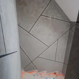 Schody w trakcie remontu, zabezpieczone pomarańczową taśmą, ściany wykończone szarą strukturą z czarnymi, geometrycznymi liniami.