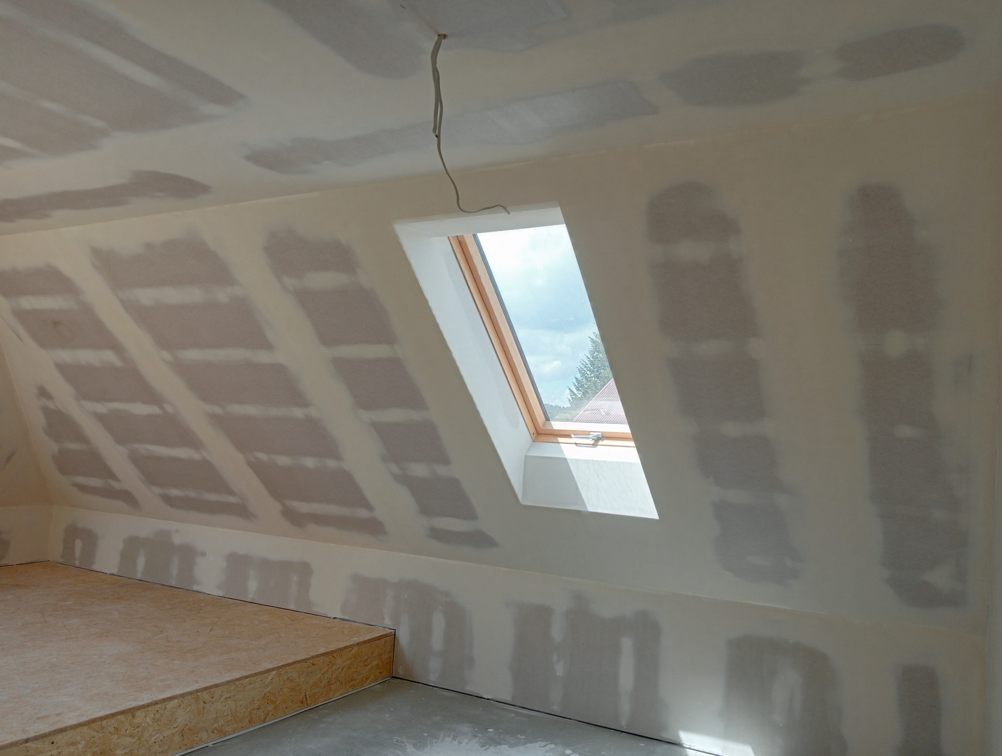 Poddasze w trakcie ocieplania, widoczne płyty gipsowo-kartonowe z widocznymi łączeniami oraz okno dachowe w drewnianej ramie z widokiem na zewnątrz.