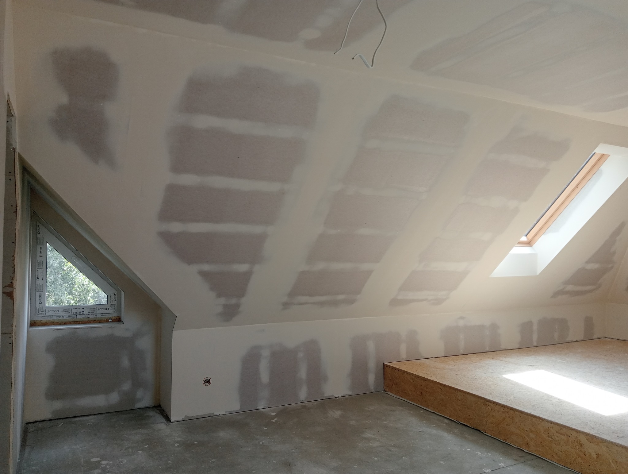 Poddasze w trakcie prac wykończeniowych: ściany pokryte gładzią szpachlową, widoczne okna połaciowe i szczytowe, przygotowana podłoga z płyt OSB.