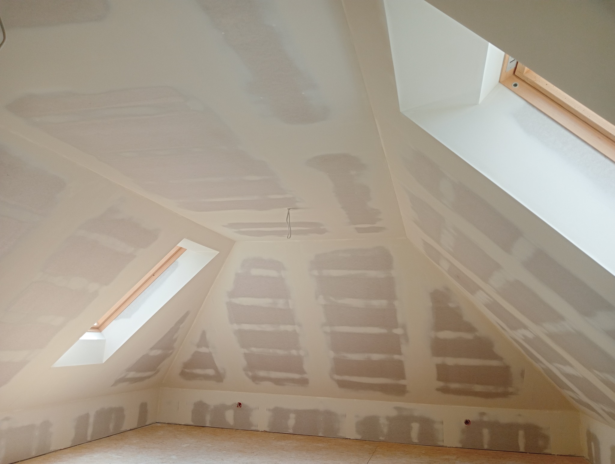 Poddasze ze skośnymi ścianami i oknami dachowymi w trakcie szpachlowania i przygotowywania do malowania, widoczne ślady po szpachli na płytach gipsowo-kartonowych.