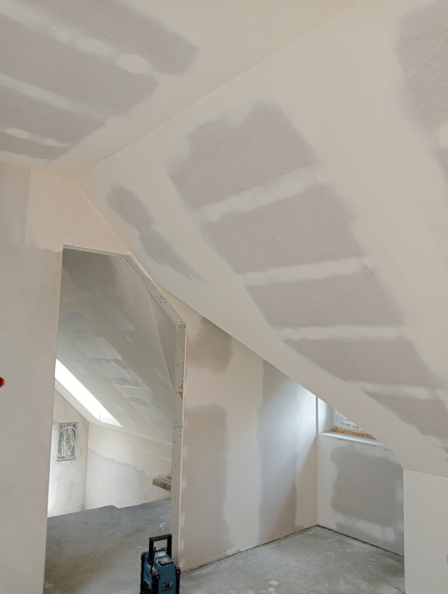 Poddasze z oknem dachowym po szpachlowaniu płyt kartonowo-gipsowych, widoczne łączenia płyt przygotowane do malowania, na podłodze laser krzyżowy.