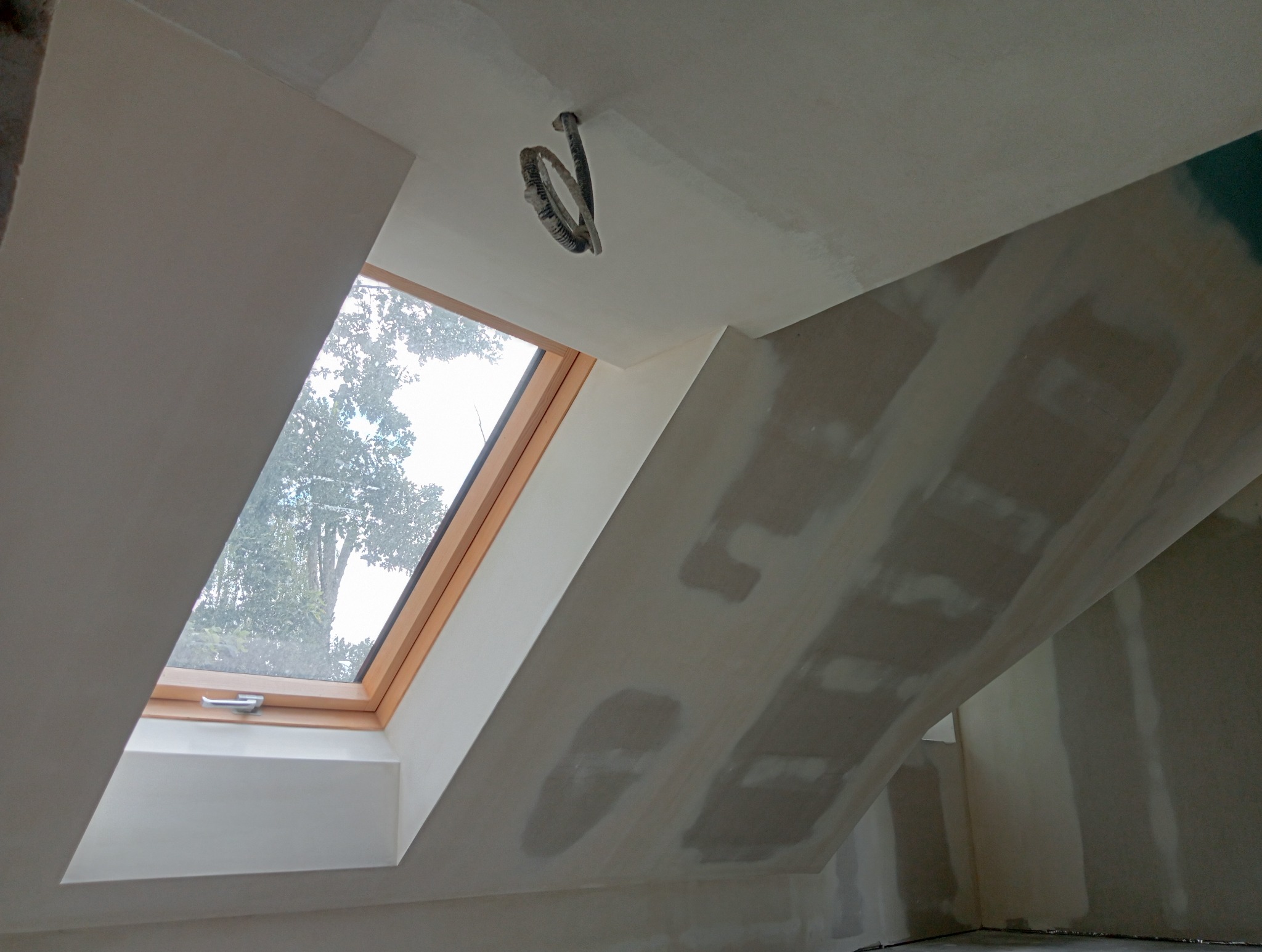 Poddasze z oknem dachowym w trakcie szpachlowania, widoczne ślady po łatach gipsowych na skosach i suficie, oraz przewody elektryczne wystające z sufitu.