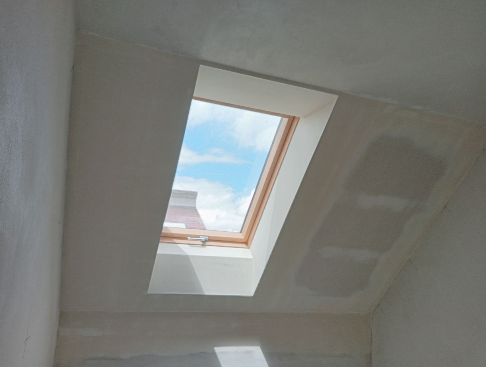 Okno dachowe z widokiem na błękitne niebo i chmury, zamontowane w ścianie poddasza w trakcie wykańczania, widoczne ślady po szpachlowaniu.