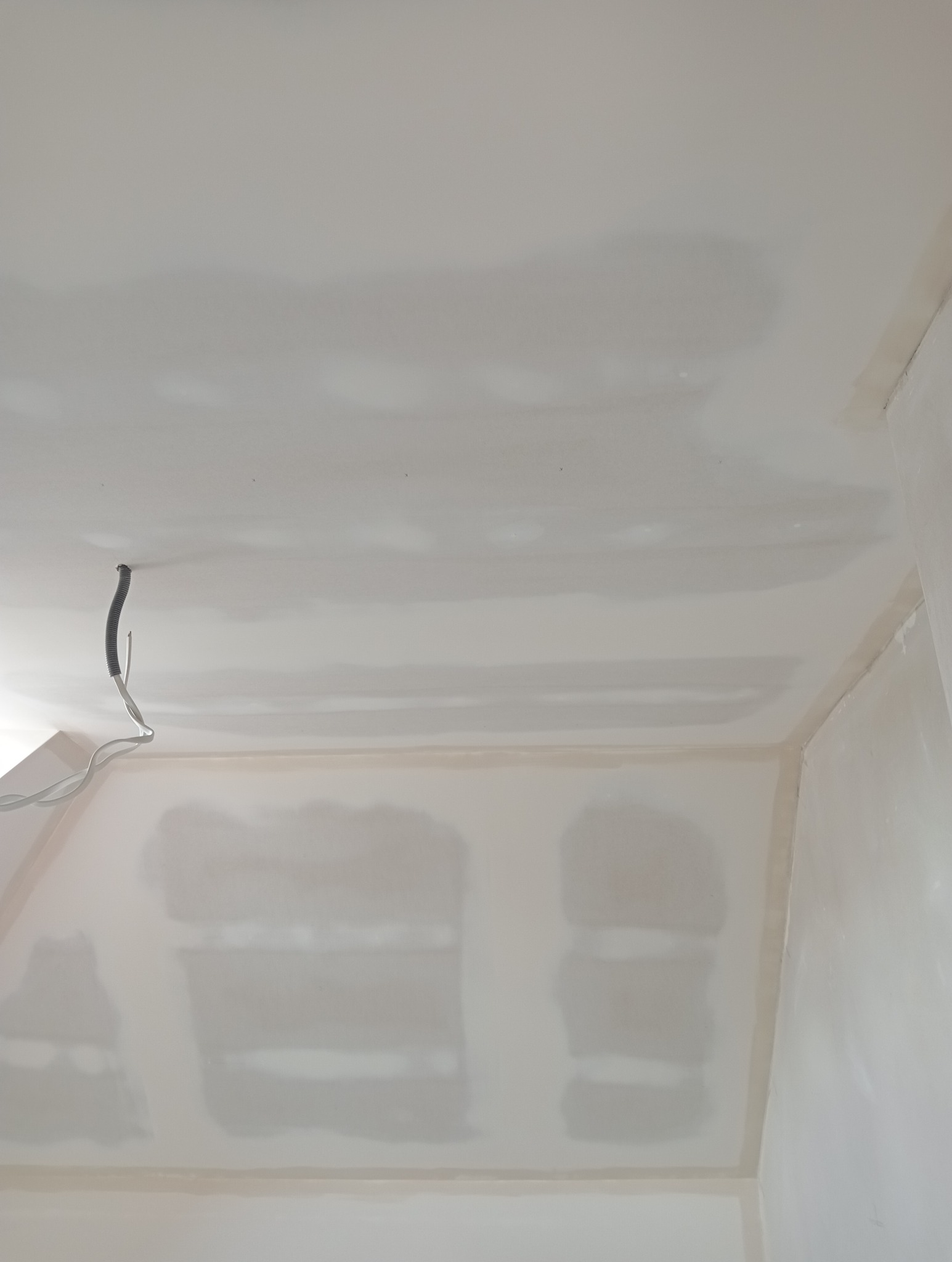 Wnętrze pokoju w trakcie wykańczania, widoczne ściany i sufit pokryte gładzią gipsową, z wystającymi przewodami elektrycznymi ze ściany.