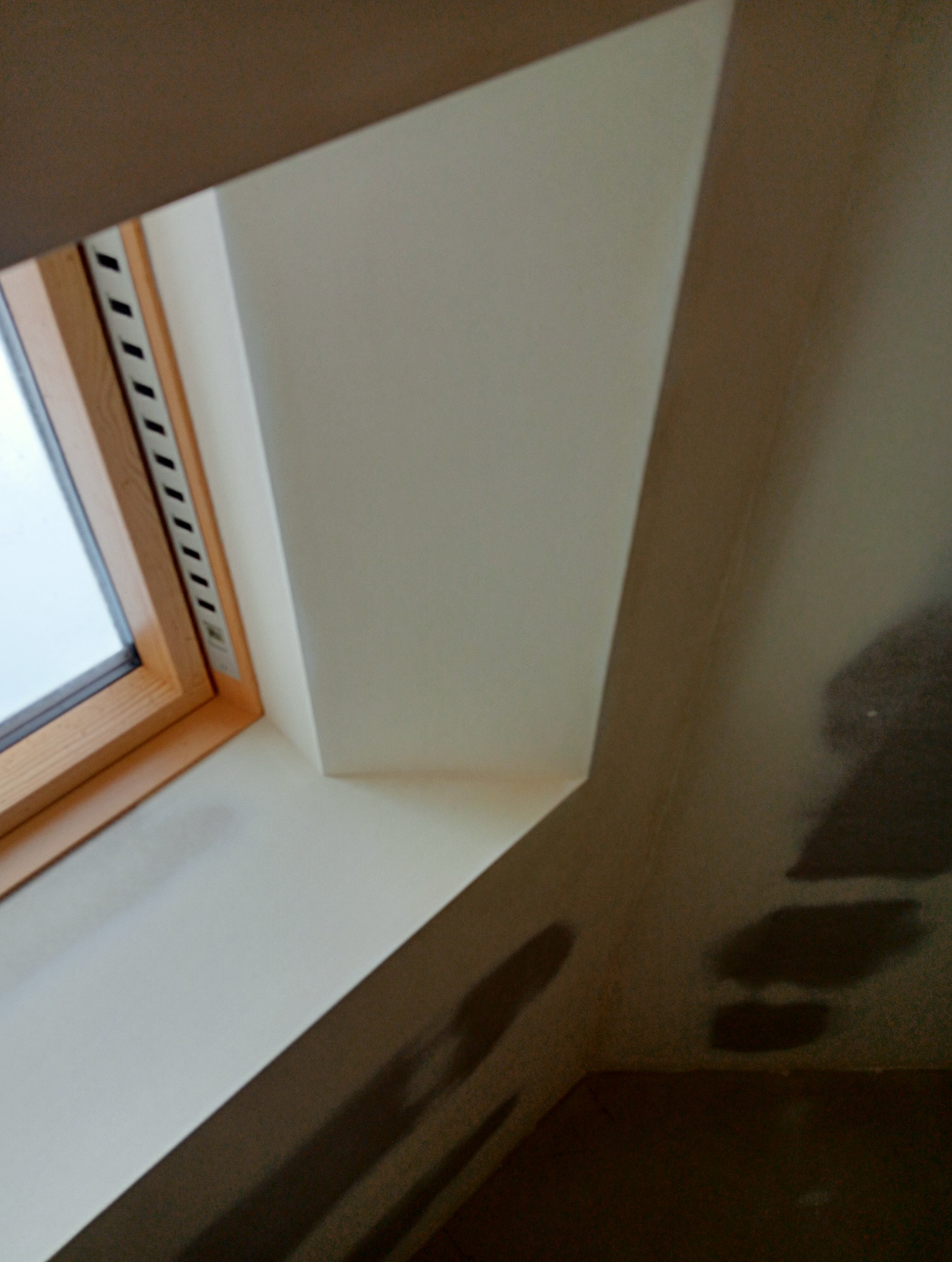 Fragment poddasza z oknem dachowym w drewnianej ramie, świeżo pomalowane na biało, widoczne niedociągnięcia malarskie na ścianie przy podłodze, ciemna plama cienia.