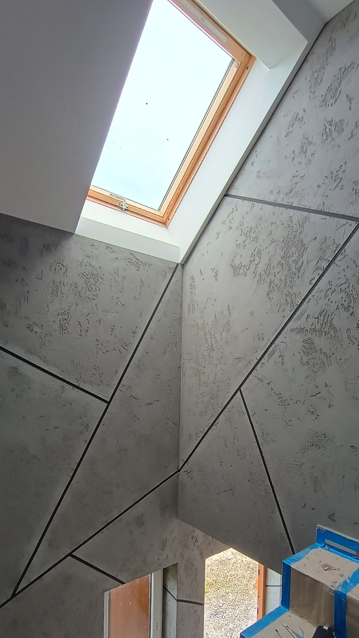 Ściana w szarym odcieniu z dekoracyjną strukturą i geometrycznym podziałem czarnymi liniami, oświetlona przez okno dachowe z drewnianą ramą, widoczny fragment schodów zabezpieczonych folią malarską.