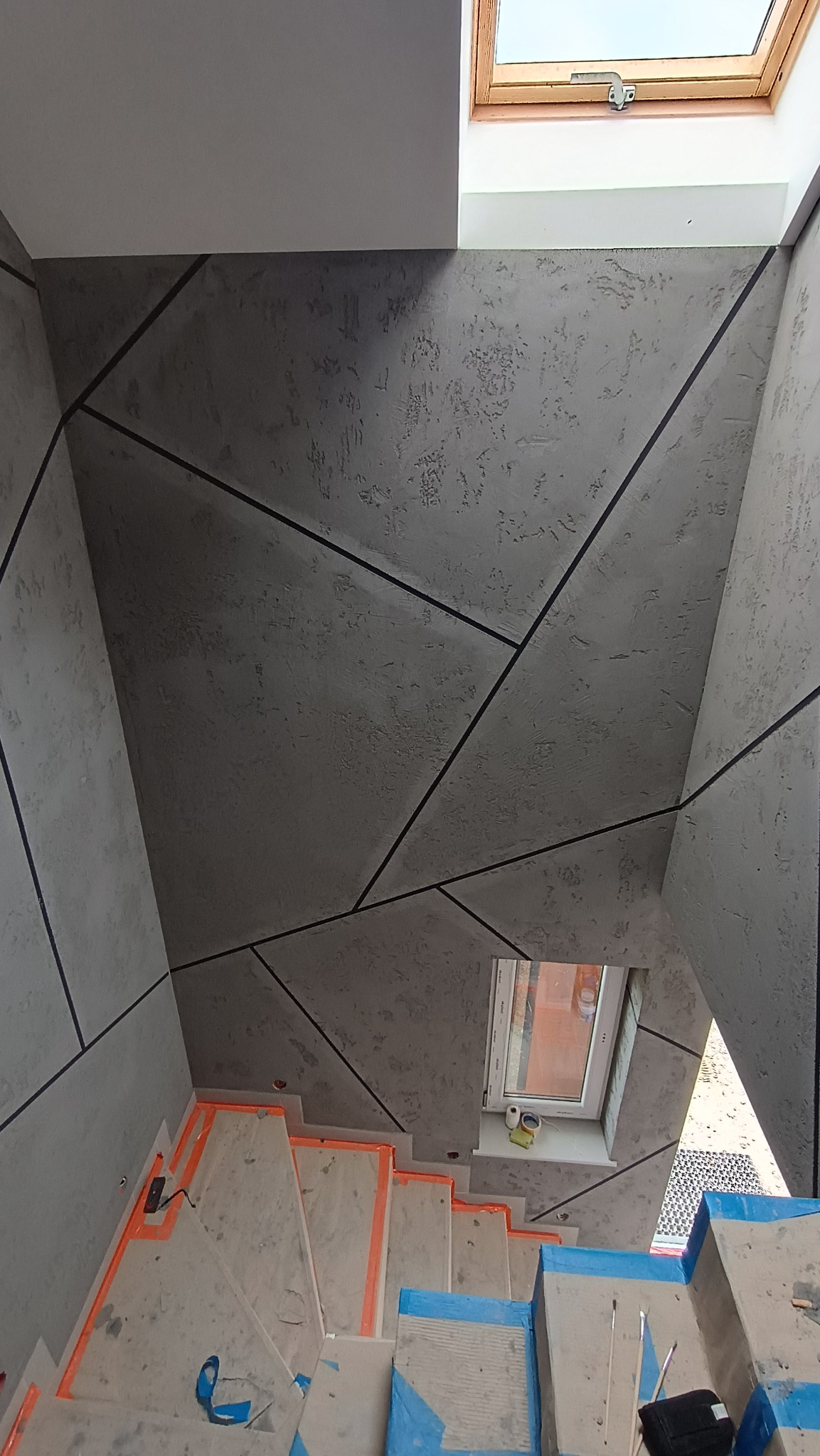 Ściany i sufit wykończone szarą, strukturalną farbą z geometrycznym wzorem czarnych linii, widok na schody zabezpieczone folią malarską i okno dachowe w trakcie prac wykończeniowych.