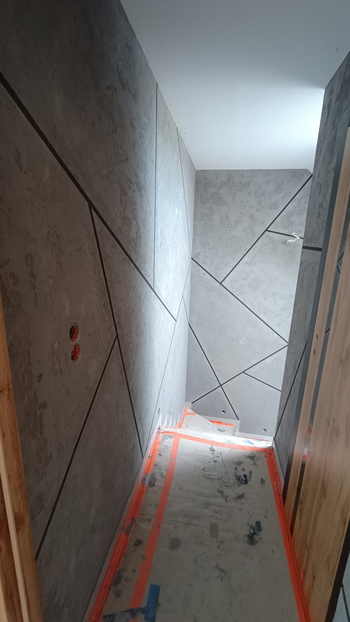 Wnętrze korytarza w trakcie wykańczania ścian dekoracyjną szarą farbą ze wzorem geometrycznym, zabezpieczona podłoga pomarańczową taśmą malarską, widoczne puszki elektryczne.