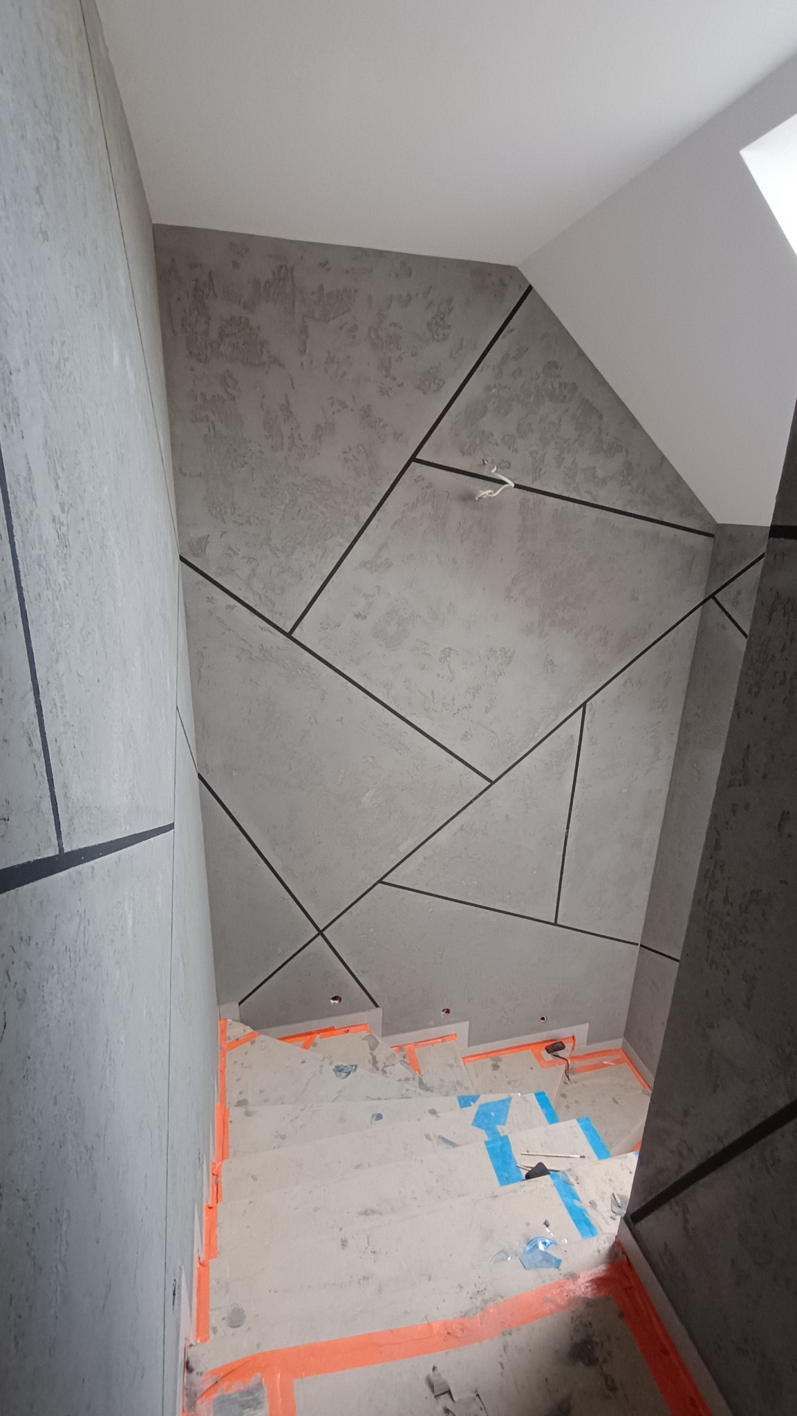 Schody w trakcie remontu, zabezpieczone pomarańczową taśmą, ściany wykończone szarą strukturą z czarnymi, geometrycznymi liniami.