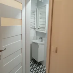 Widok na małą, odnowioną łazienkę przez uchylone białe drzwi z poziomymi wgłębieniami, ukazujący umywalkę na szafce, grzejnik drabinkowy i czarno-białą wzorzystą podłogę.