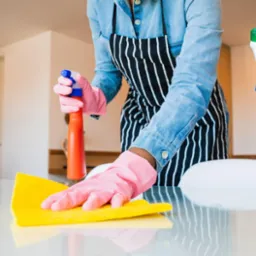Osoba w różowych rękawicach i fartuchu w paski czyści blat żółtą ścierką i pomarańczowym sprayem, z boku widoczne wiadro z detergentami.