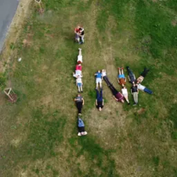 Widok z drona na grupę osób leżących na trawie, tworzących kształt litery 'K', z parą obejmującą się w tle.