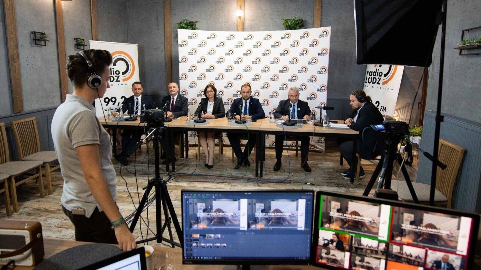 Kobieta w słuchawkach kontroluje nagranie z panelu dyskusyjnego, widocznego na monitorach; w tle grupa osób siedzi za stołem z mikrofonami, przed ścianą z logo Radio Łódź.