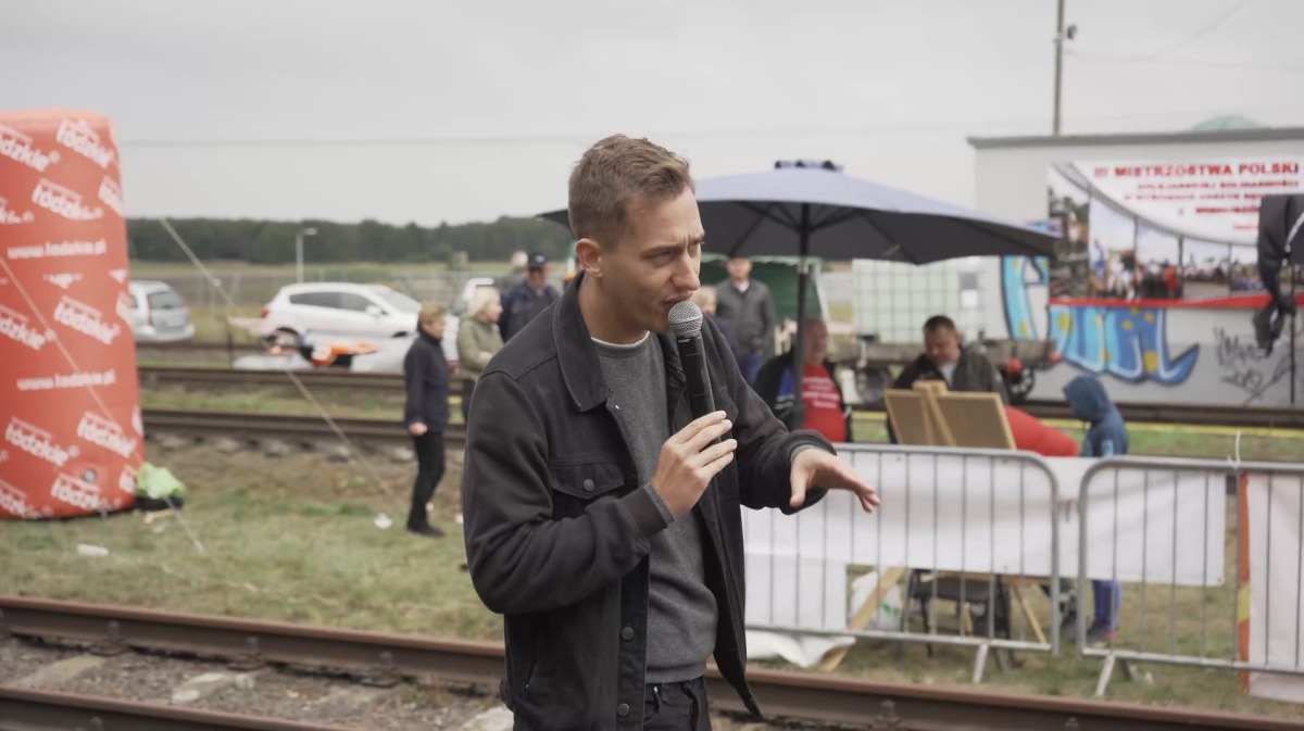Mężczyzna z mikrofonem przemawia na tle sceny z napisem 'Mistrzostwa Polski', widoczny dmuchany baner reklamowy i grupa osób.