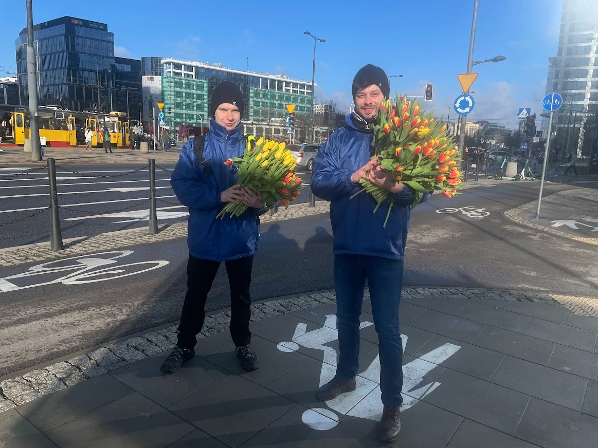 Dwaj mężczyźni w niebieskich kurtkach trzymają bukiety tulipanów na tle miejskiego krajobrazu z tramwajem i nowoczesnymi budynkami.