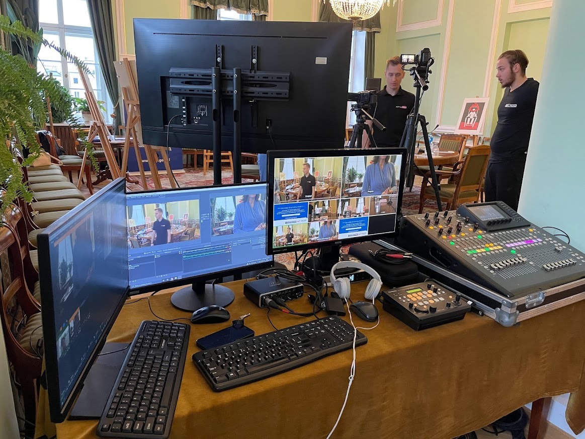 Stanowisko do realizacji transmisji na żywo z dwoma operatorami, kamerą na statywie, mikserem audio i podglądem na monitorach w eleganckim wnętrzu z żyrandolem.