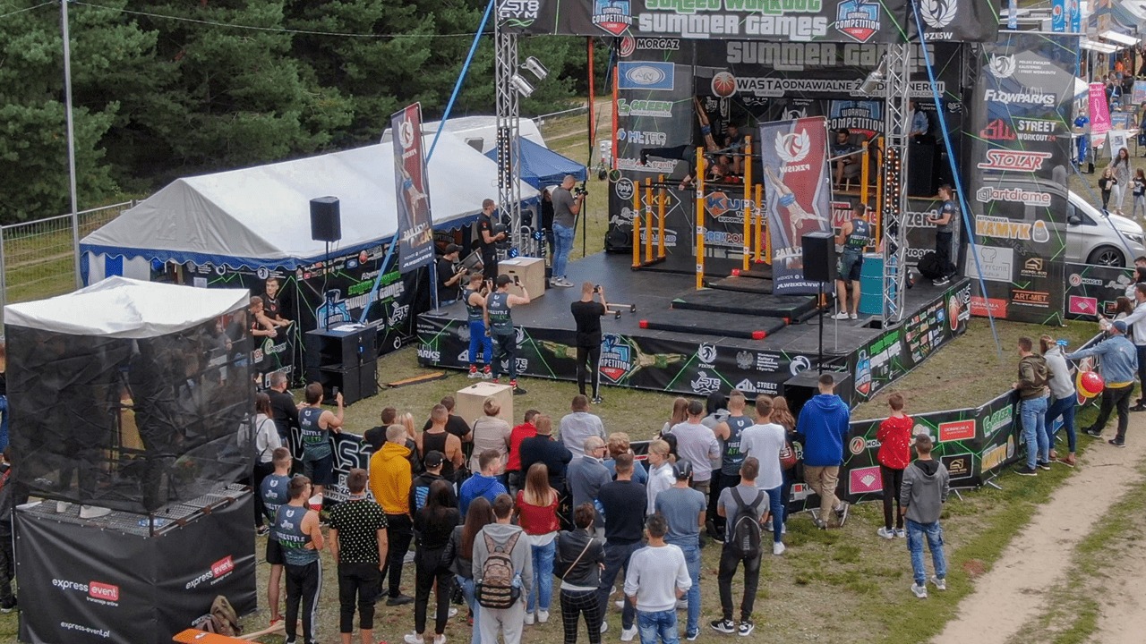 Widok z góry na plenerową scenę podczas zawodów Street Workout Summer Games, z uczestnikami prezentującymi akrobacje na drążkach, fotografami dokumentującymi wydarzenie, publicznością obserwującą...