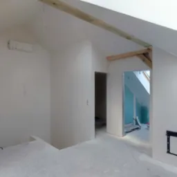 Poddasze w trakcie wykańczania: białe ściany po gładzi, widoczne drewniane elementy konstrukcyjne, przygotowane otwory drzwiowe, instalacja elektryczna w ścianie, fragment ściany z płyty karton...