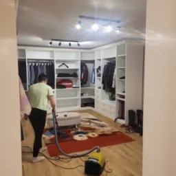 Osoba odkurza wzorzysty dywan w garderobie z białymi szafami, widoczny puf i buty.