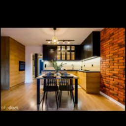 Nowoczesna, jasna kuchnia z drewnianymi szafkami, czarnymi elementami i ceglaną ścianą. Stół z czarnymi krzesłami, nakryty do posiłku.