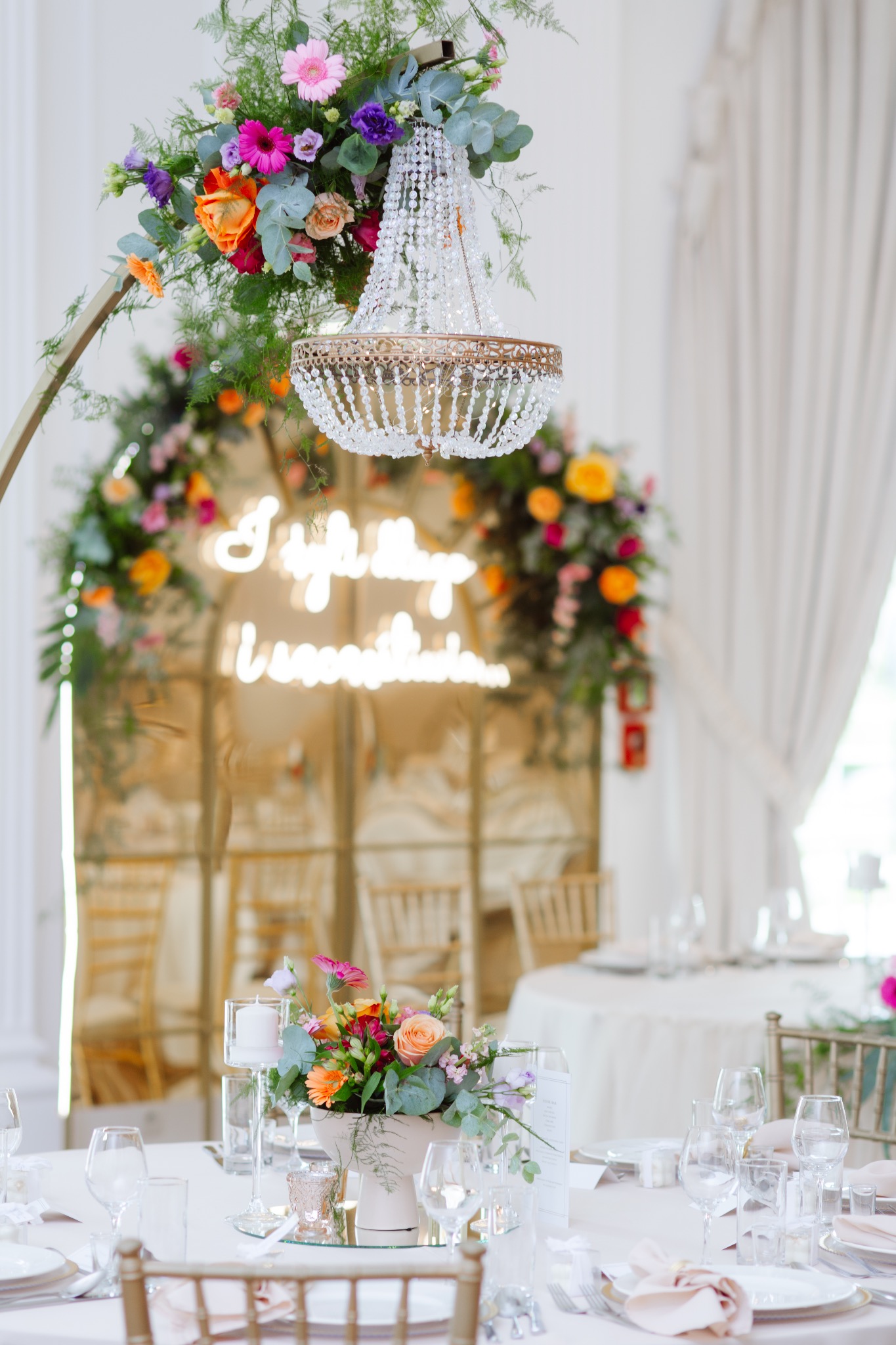 Uroczysta aranżacja stołu weselnego z kwiatami w odcieniach różu, pomarańczy i fioletu, kryształowym żyrandolem i neonowym napisem w tle.