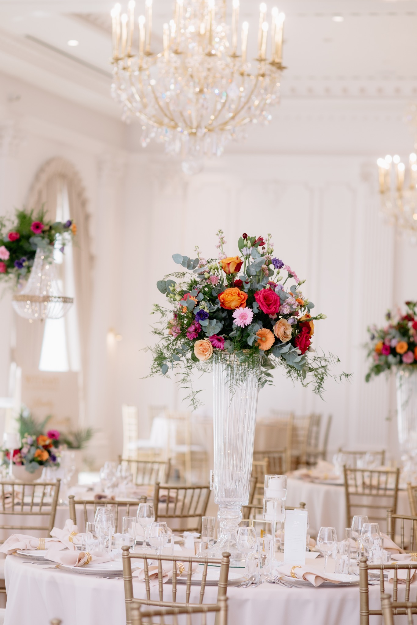 Uroczysta dekoracja stołu weselnego z wysokim, przezroczystym wazonem wypełnionym kolorowymi kwiatami, złotymi krzesłami i kryształowymi żyrandolami w tle.