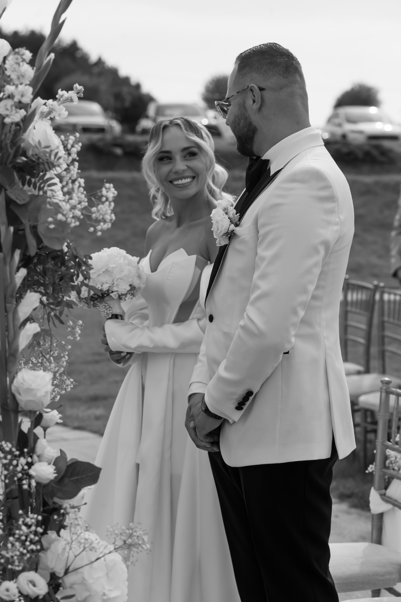 Czarno-białe zdjęcie uśmiechniętej panny młodej w białej sukni z bukietem, stojącej obok pana młodego w białym smokingu, na tle dekoracji kwiatowych i samochodów.