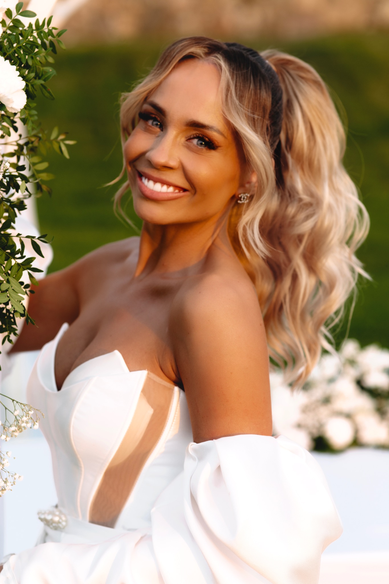 Uśmiechnięta blondynka w białej sukni z bufiastymi rękawami, pozująca przy białej konstrukcji z kwiatami i zielenią, w tle rozmyta zieleń.