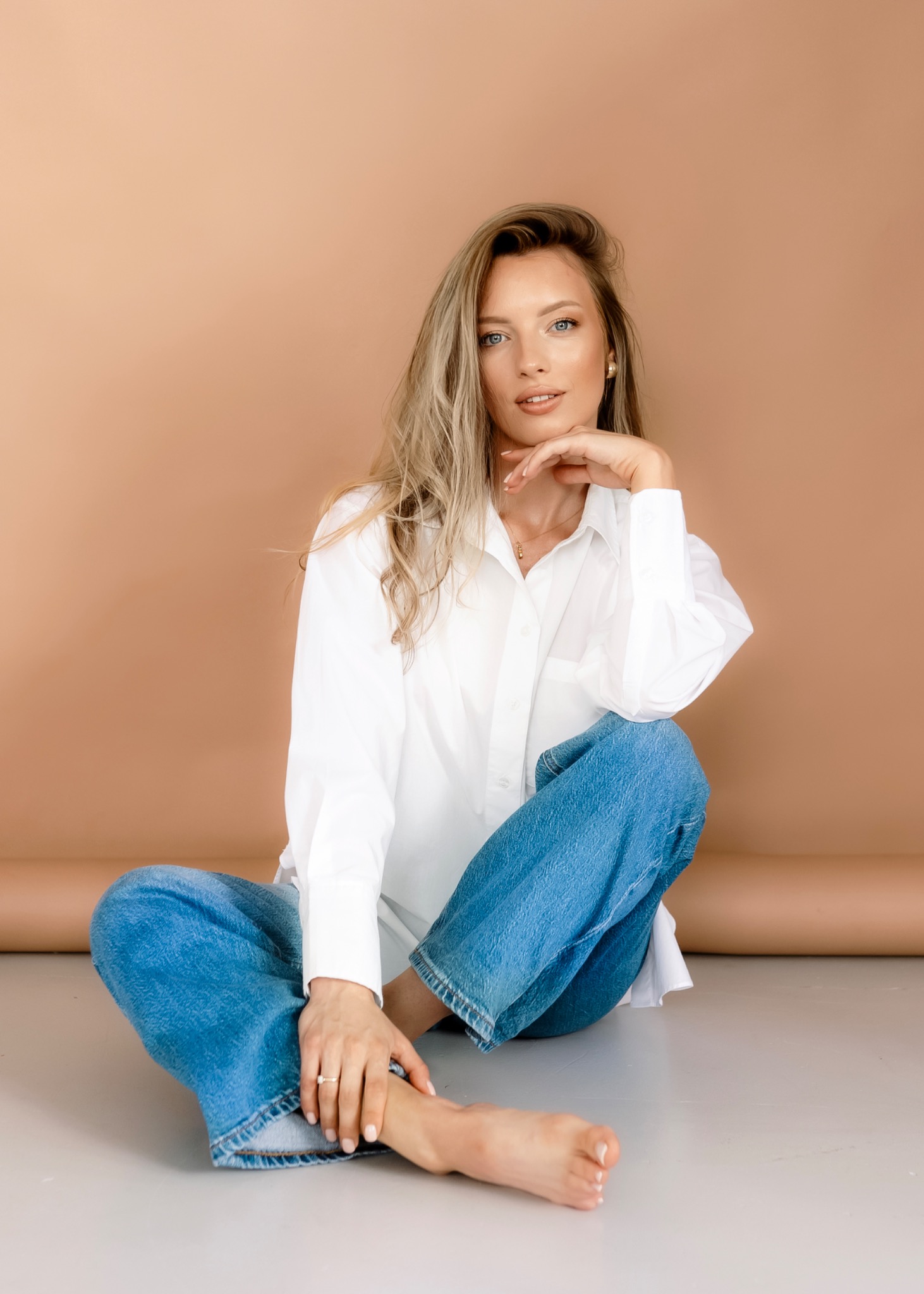 Portret kobiety siedzącej na podłodze w białej koszuli i jeansach, z delikatnym makijażem i złotą biżuterią na beżowym tle.