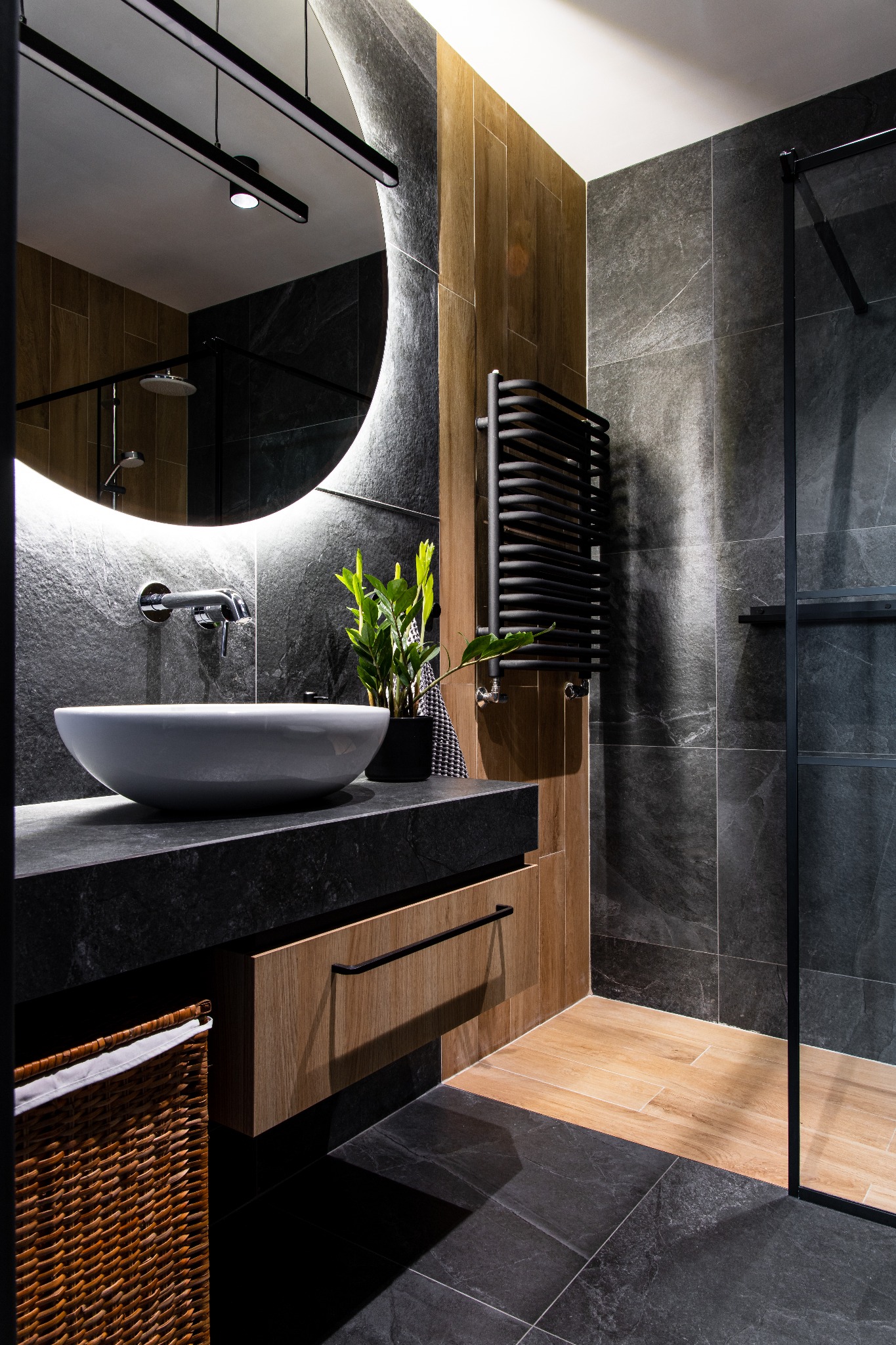 Nowoczesna łazienka z okrągłym lustrem podświetlanym LED, umywalką nablatową, czarnym grzejnikiem drabinkowym i kabiną prysznicową typu walk-in z czarnymi profilami.