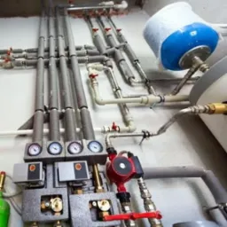 Rozbudowana instalacja hydrauliczna z licznymi rurami, zaworami, manometrami i pompą obiegową, zamontowana przy białej ścianie.