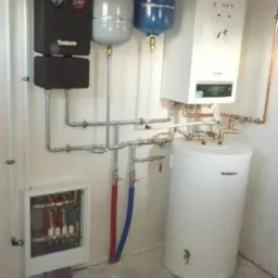 Nowoczesna kotłownia z kotłem gazowym, zbiornikiem CWU, naczyniami wzbiorczymi i rozdzielaczem, wszystko marki Buderus, zamontowana w surowym pomieszczeniu.