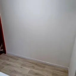 Pomalowana na biało ściana i ułożone panele podłogowe w jasnym odcieniu drewna, widoczny fragment sprzętu AGD w rogu.