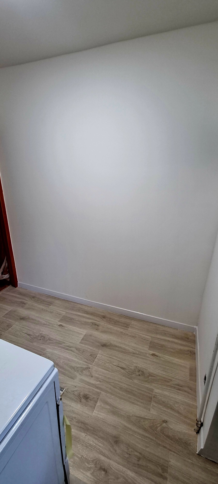 Pomalowana na biało ściana i ułożone panele podłogowe w jasnym odcieniu drewna, widoczny fragment sprzętu AGD w rogu.