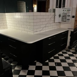 Daniel Jesionkowski - Czarne szafki z białym blatem i białymi kafelkami na ścianie, uzupełnione skrzynką elektryczną, na podłodze czarno-biała szachownica.