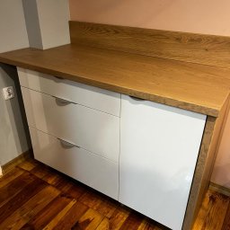 Daniel Jesionkowski - Nowoczesna komoda z trzema białymi szufladami i blatem z jasnego drewna, stojąca na drewnianej podłodze, widoczny fragment ściany w dwóch odcieniach.