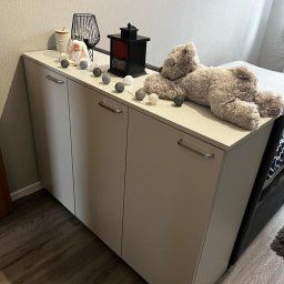 Daniel Jesionkowski - Trzydrzwiowa szafka z szarymi frontami i podłużnymi uchwytami, udekorowana lampkami cotton balls, lampionem i pluszowym misiem leżącym na blacie, częściowo zasłaniającym łóżko.