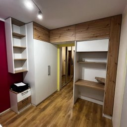 Daniel Jesionkowski - Biała zabudowa meblowa z elementami drewnopodobnymi w pokoju, obejmująca szafę z czarnymi uchwytami, regał z półkami i biurko z półkami, oświetlona lampą sufitową na szynie.