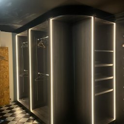 Daniel Jesionkowski - Czarna, narożna garderoba z oświetleniem LED, widoczne półki i drążki na wieszaki, na podłodze czarno-biała szachownica.